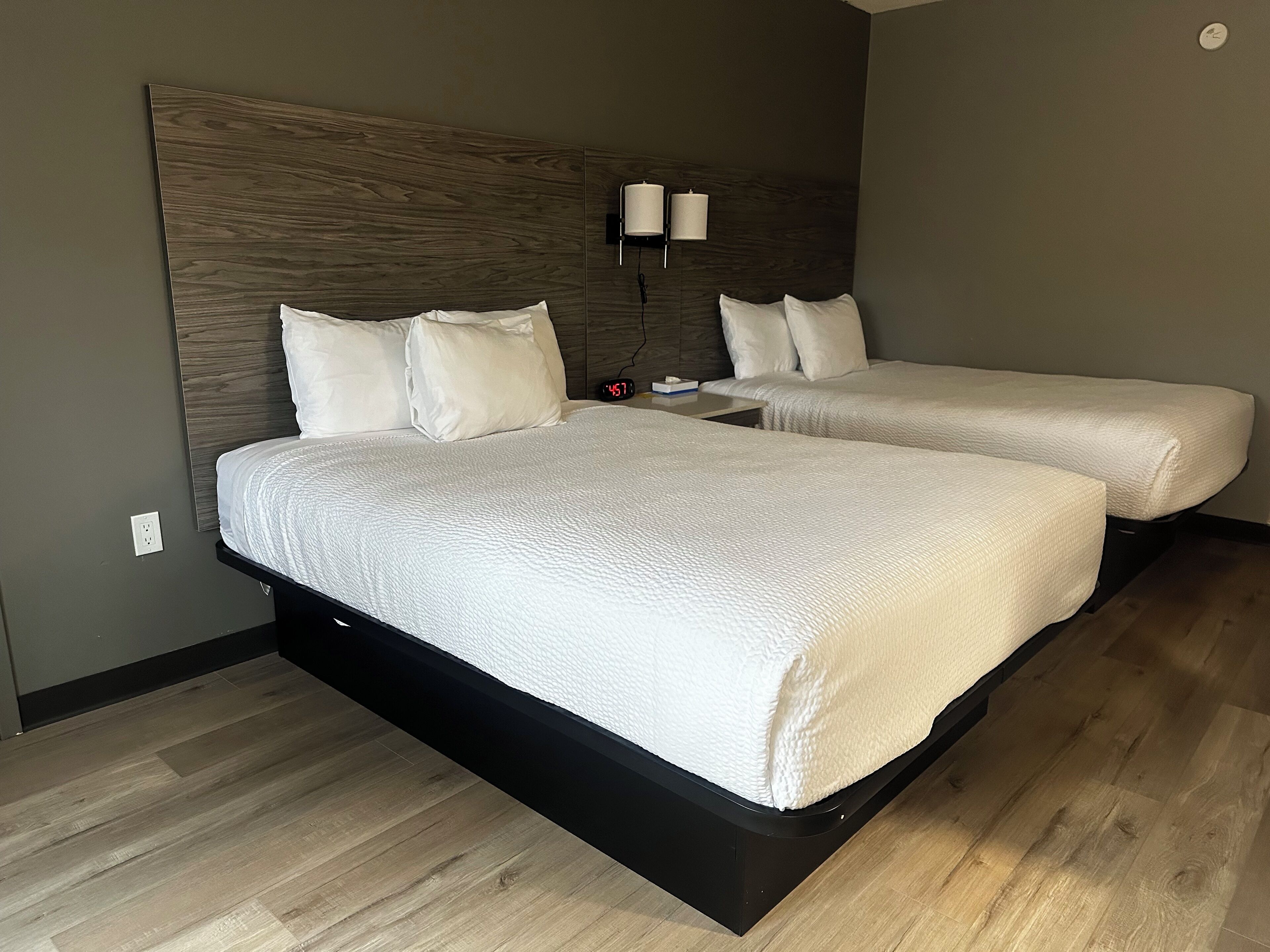 Une chambre d’hôtel avec deux lits, une tête de lit en bois, une table de chevet avec une lampe et une horloge.