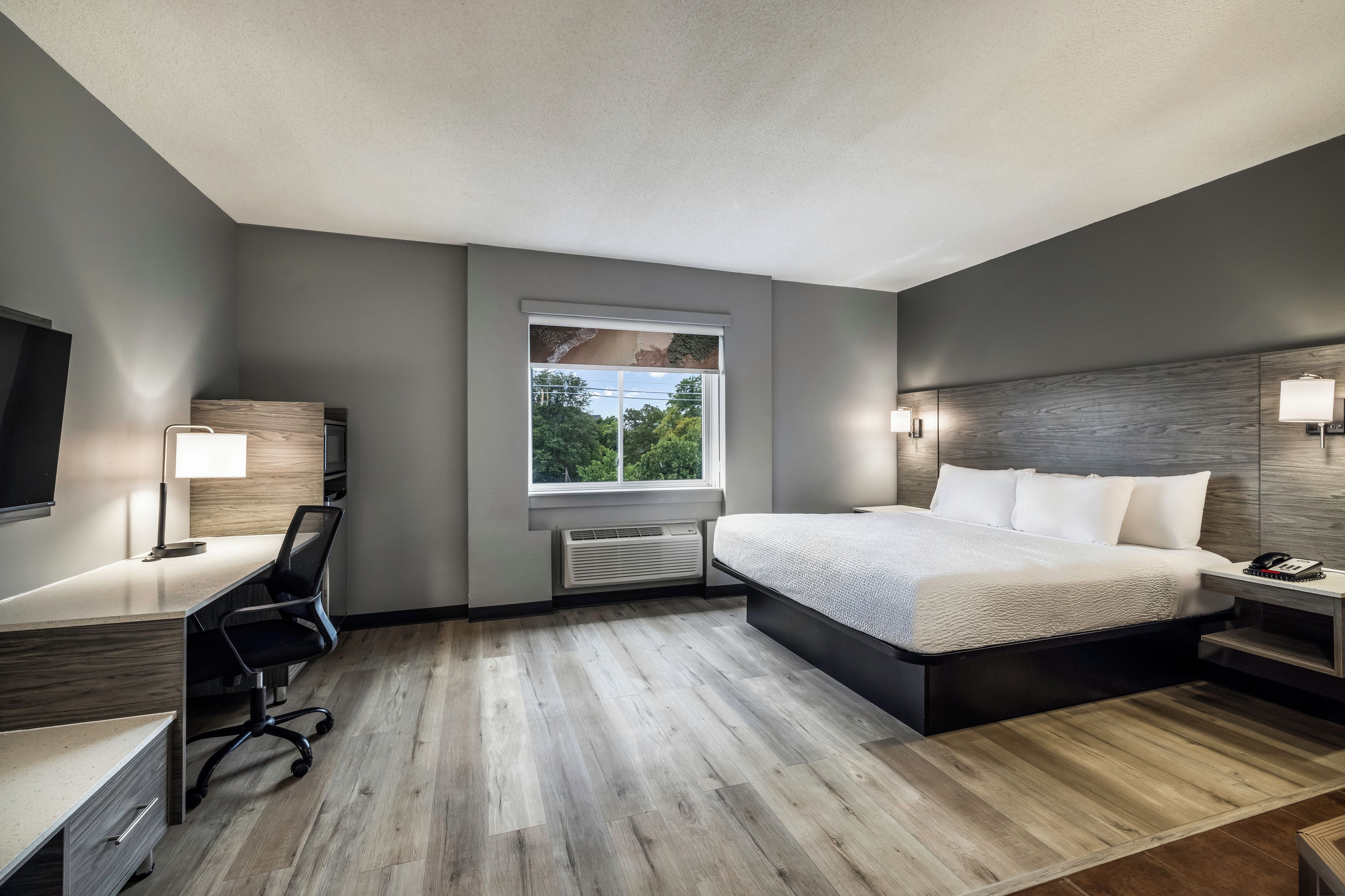 A modern hotel room with a bed, desk, and chair, a large window, and a flat-screen TV.
