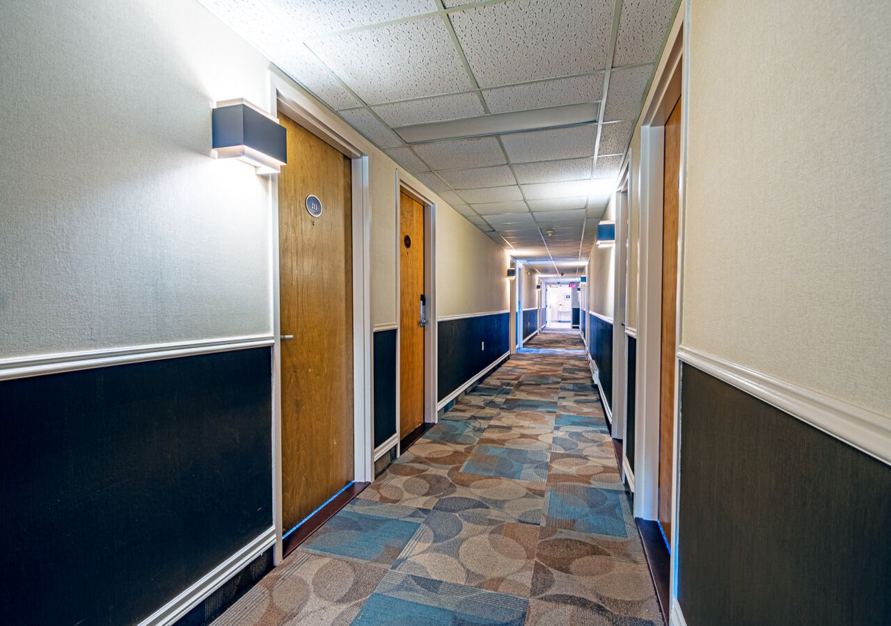 A long, narrow hotel corridor with patterned carpet, wooden doors, and a ceiling with fluorescent lighting.