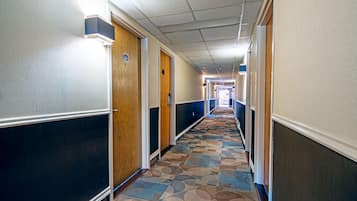 A long, narrow hotel corridor with patterned carpet, wooden doors, and a ceiling with fluorescent lighting.