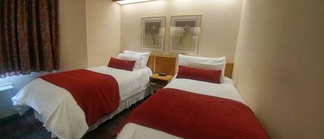 Habitación de hotel con dos camas, cada una con ropa de cama blanca y detalles rojos, una mesita de noche y dos cuadros con motivos florales.