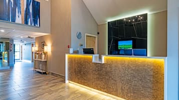 A hotel reception area with a wooden reception desk, a mounted television, and a large abstract painting on the wall.