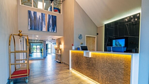 A hotel reception area with a wooden reception desk, a gold cart, and a large window with a view of the lobby. - Slumber Inn (New Minas)