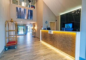 A hotel reception area with a wooden reception desk, a gold cart, and a large window with a view of the lobby.