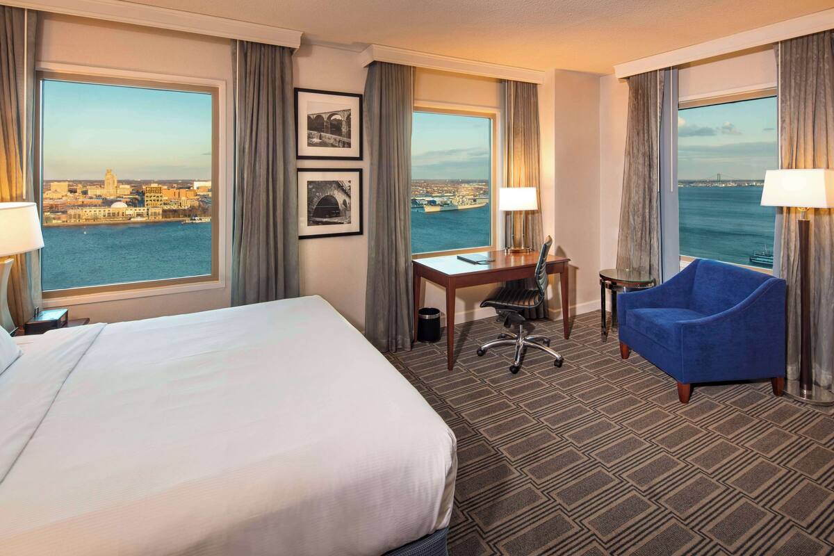 Une chambre d’hôtel avec un grand lit, un bureau avec une chaise, un fauteuil bleu et deux fenêtres donnant sur la ville et le port.