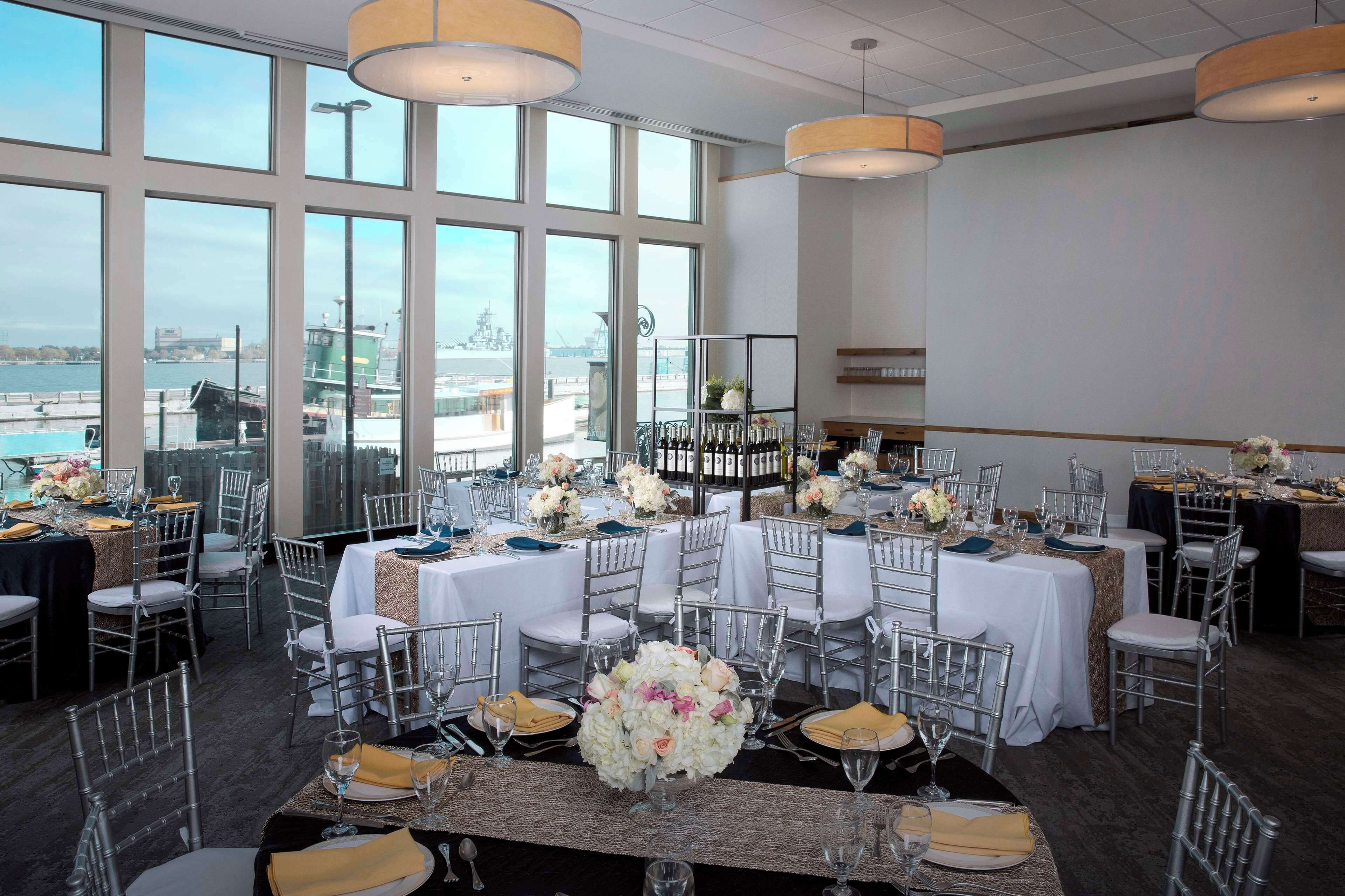 A banquet hall with multiple tables set for a formal event, large windows offering a view of the waterfront, and modern pendant lighting.