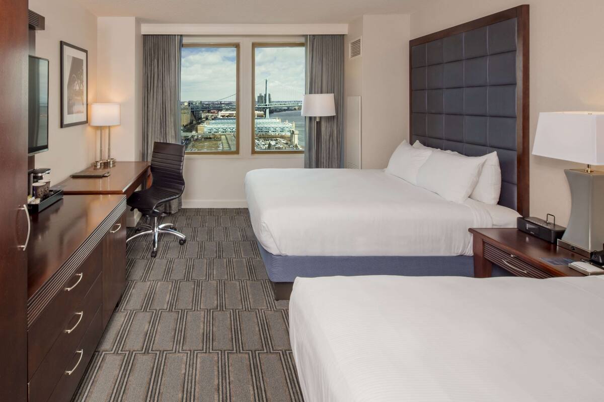 Une chambre d’hôtel avec deux lits, un bureau, une chaise et une vue sur le paysage urbain, grâce à de grandes fenêtres.