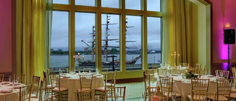 Un comedor con mesas dispuestas para una comida, ventanas grandes con cortinas y vista a un barco y al agua.