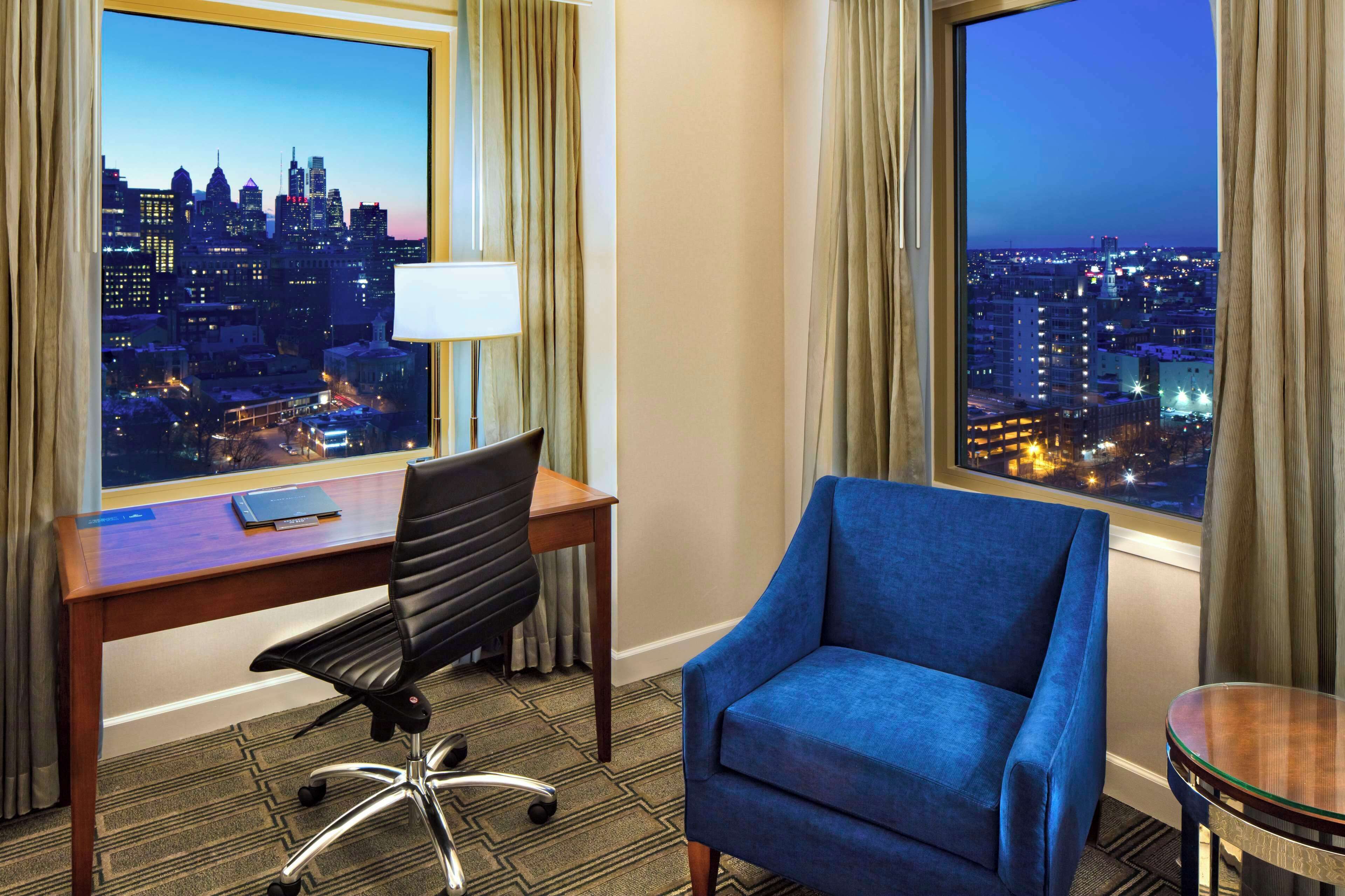 A hotel room with a desk, chair, and a large window offering a cityscape view.