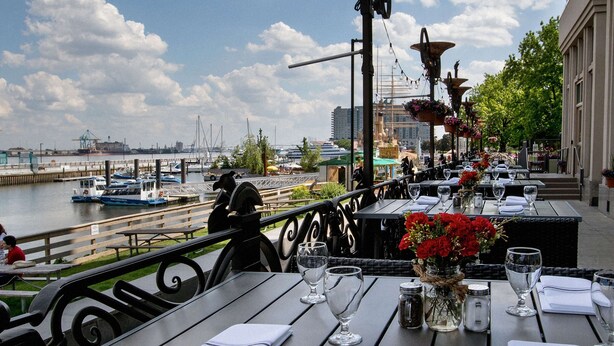 An outdoor dining area with tables set for two, overlooking a marina with boats and a dock. - Hilton Philadelphia at Penn's Landing (Philadelphia)