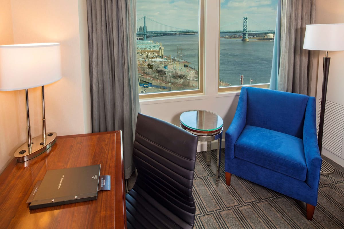 Une chambre d’hôtel avec vue sur un pont, un fauteuil bleu, une table en bois avec une lampe et un ordinateur portable.