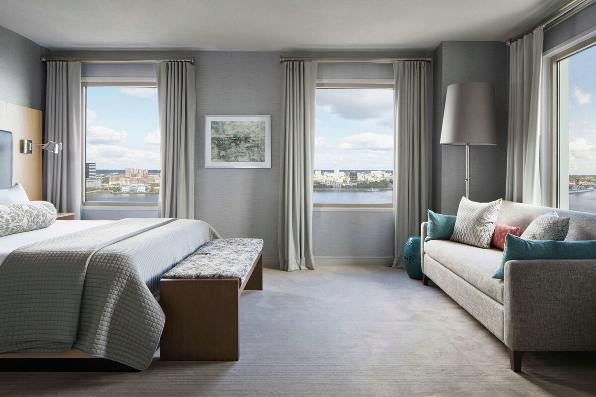 A hotel room with a bed, a sofa, a bench, a lamp, and large windows offering a city view.