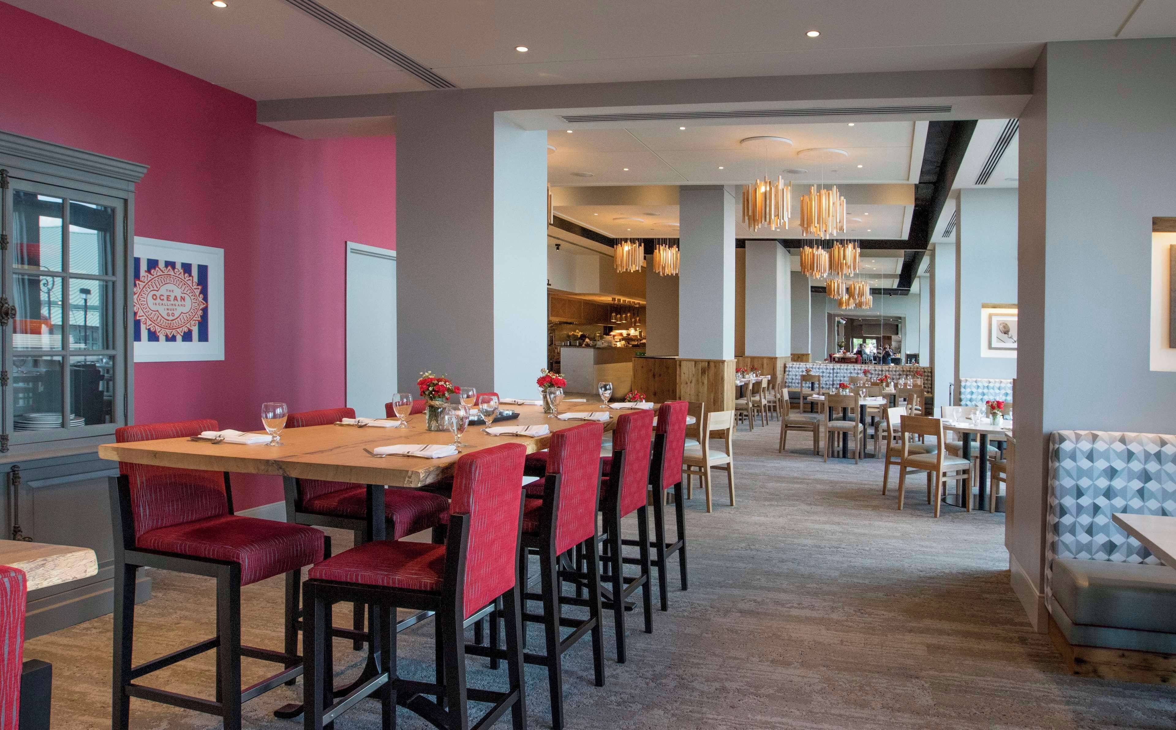 Un restaurante con una larga mesa de madera y sillas tapizadas rojas, una cocina al fondo y una zona de bar con asientos de estampado azul.