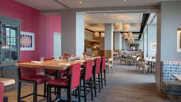 Un restaurante con una larga mesa de madera y sillas tapizadas rojas, una cocina al fondo y una zona de bar con asientos de estampado azul.
