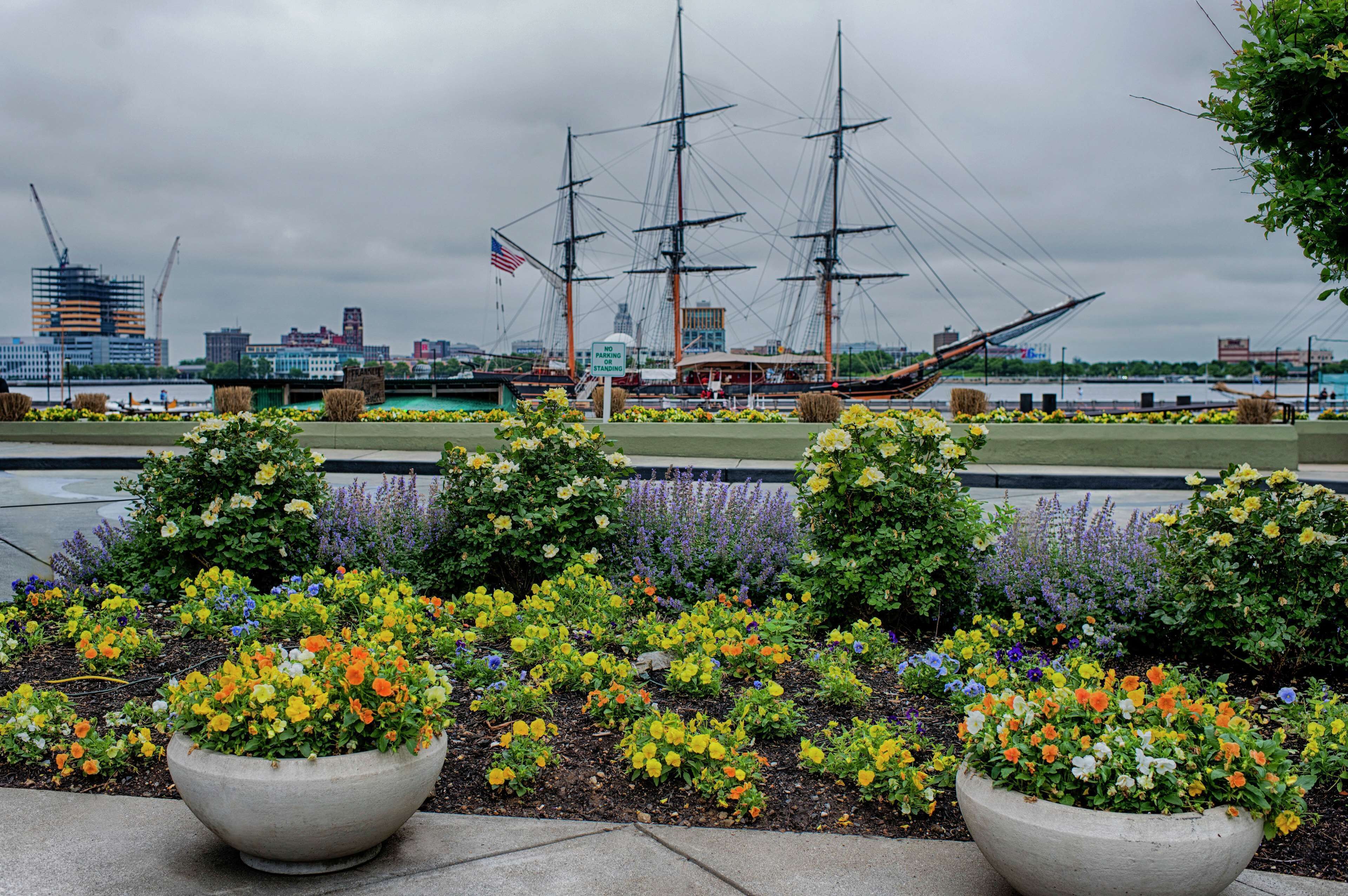Un jardín frente al agua con parterres de flores y un barco velero al fondo.