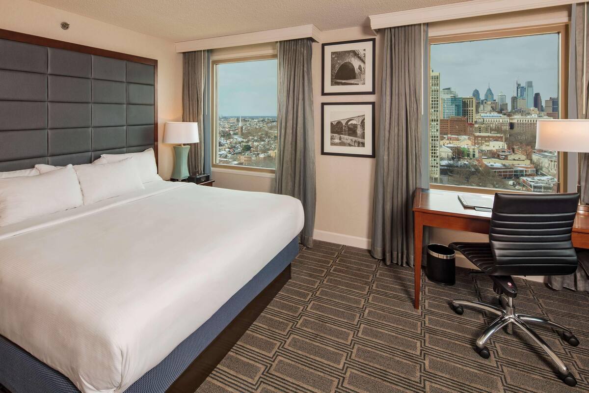 Habitación de hotel con una cama grande, un escritorio, una silla y vistas a un paisaje urbano a través de un ventanal.