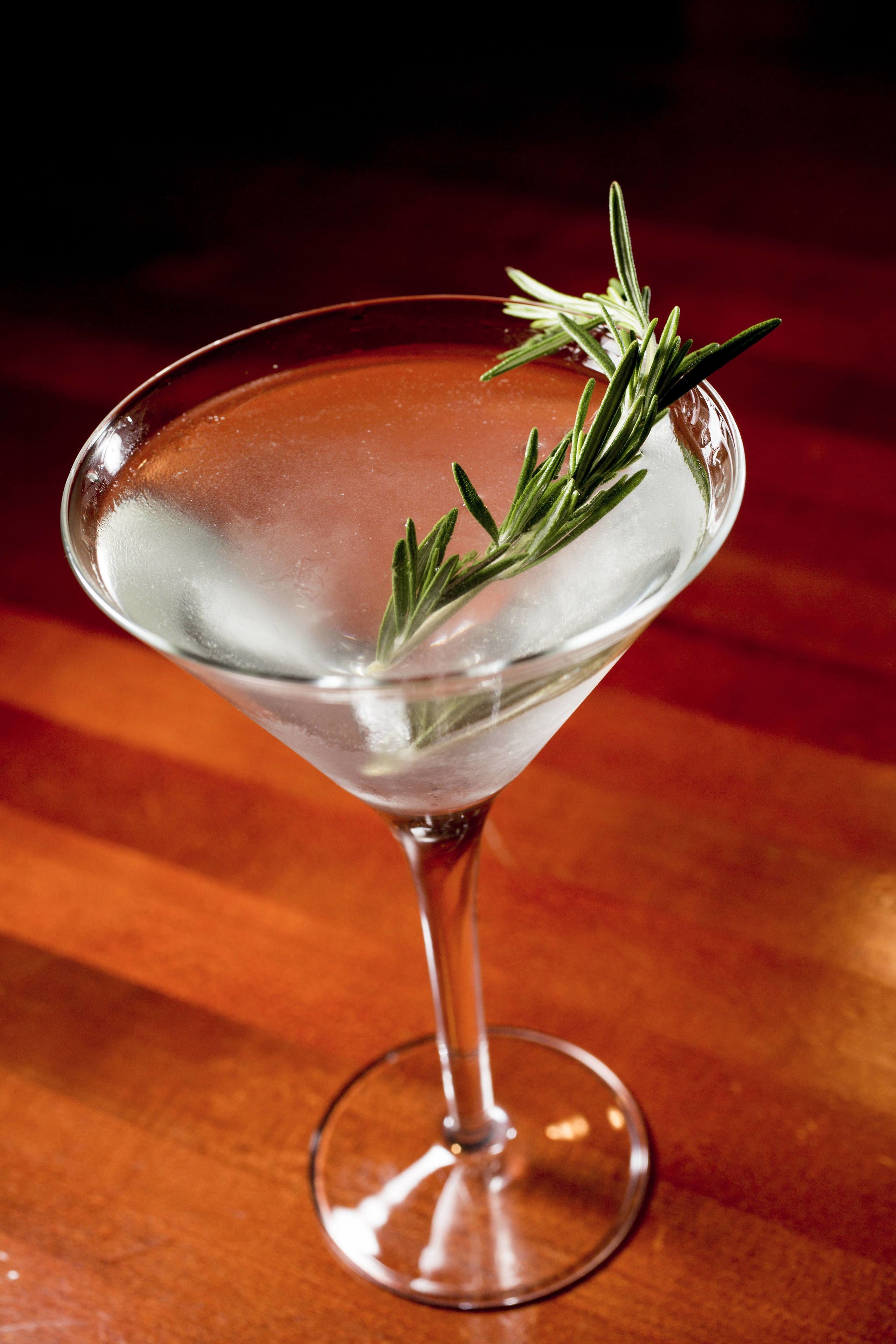 A martini glass with ice and a sprig of rosemary on a wooden surface.