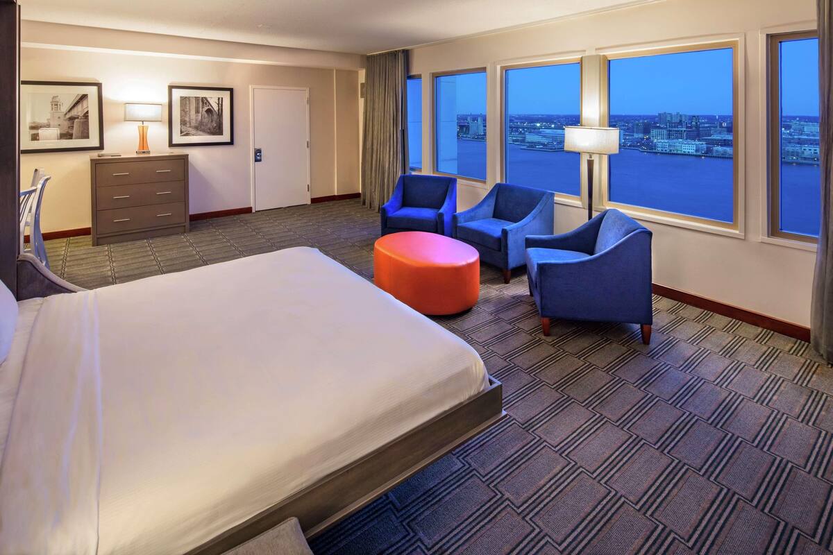 Habitación de hotel con una cama grande, dos sillones azules, un pequeño taburete naranja, un escritorio con lámpara y vistas a la ciudad a través de amplios ventanales.