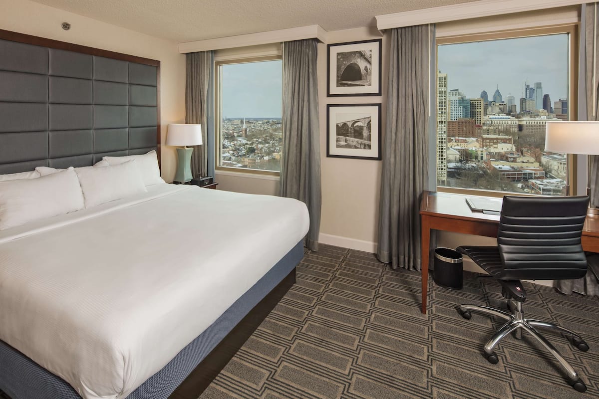 Une chambre d’hôtel avec un grand lit, un bureau avec une chaise, une fenêtre donnant sur la ville et deux tableaux encadrés au mur.