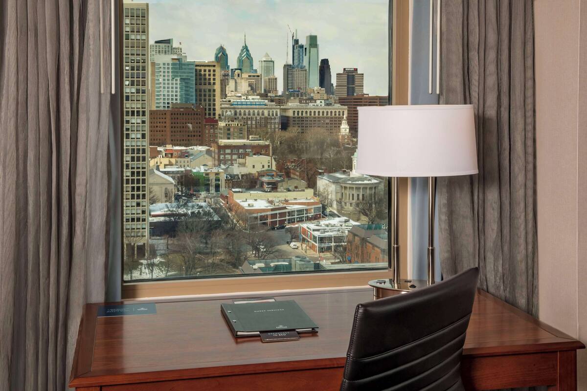 Une chambre d’hôtel équipée d’un bureau, d’une lampe et offrant une vue sur le paysage urbain grâce à une grande fenêtre.