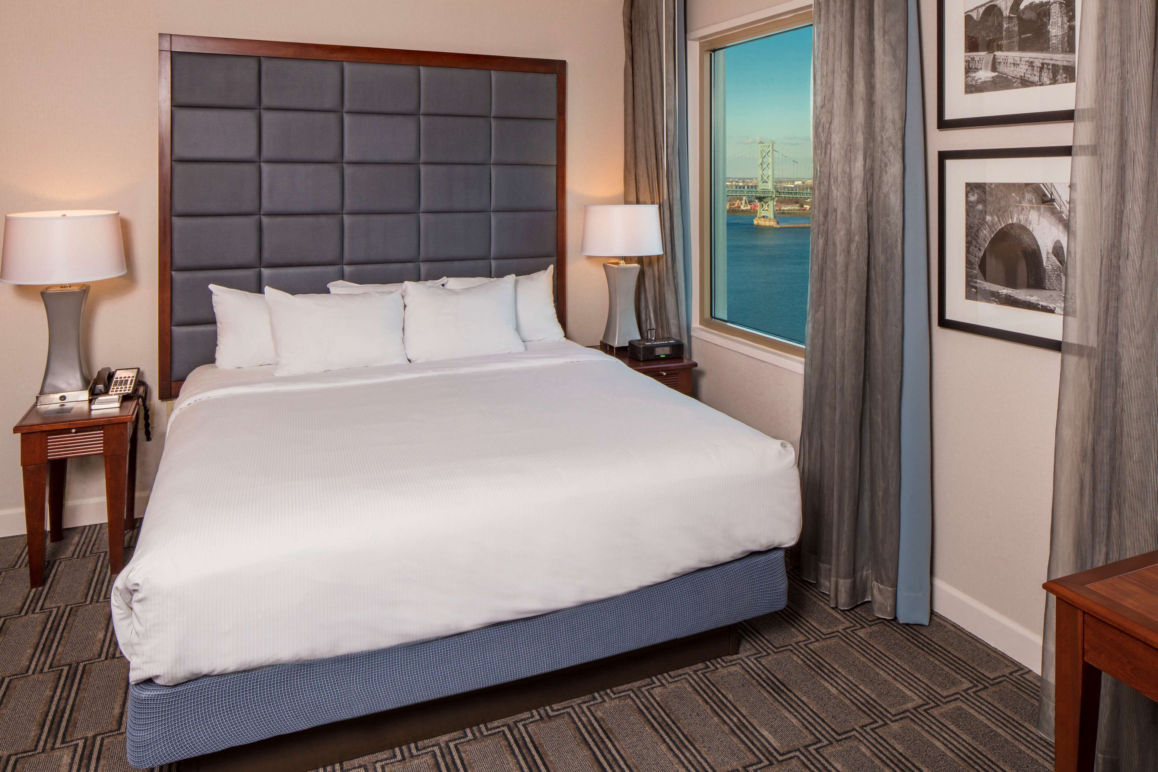 A hotel room with a large bed, two bedside tables with lamps, a view of a bridge, and framed pictures on the wall.