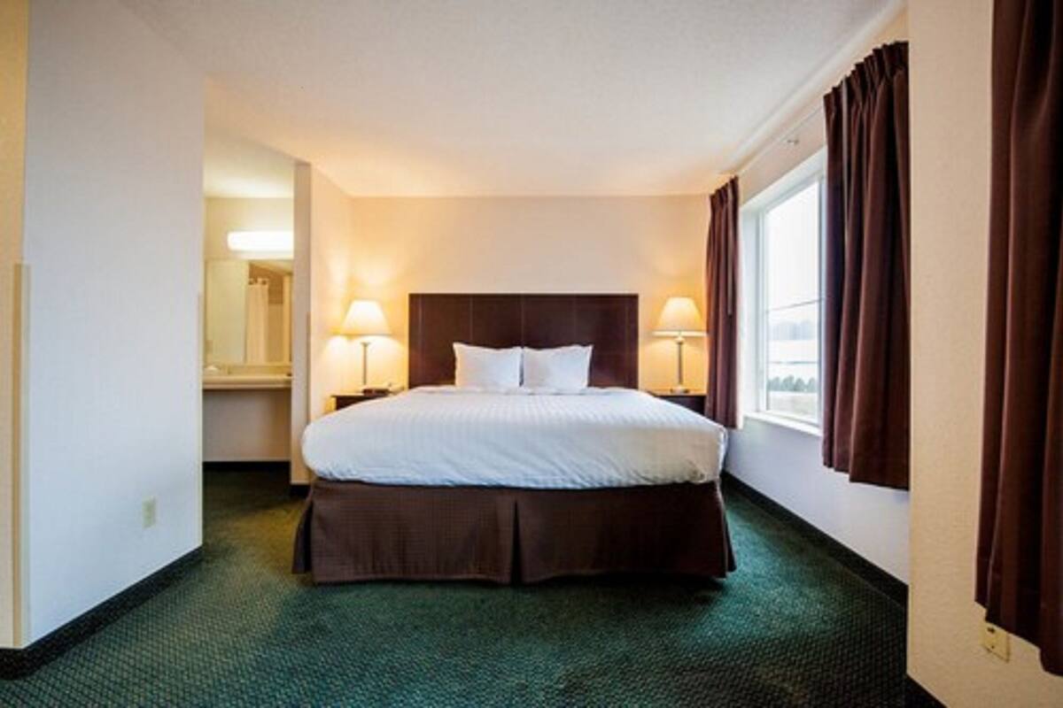 A hotel room with a large bed, two bedside lamps, a window with curtains, and a carpeted floor.