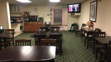 A hotel dining area with multiple tables and chairs, a coffee station, and a television on the wall.