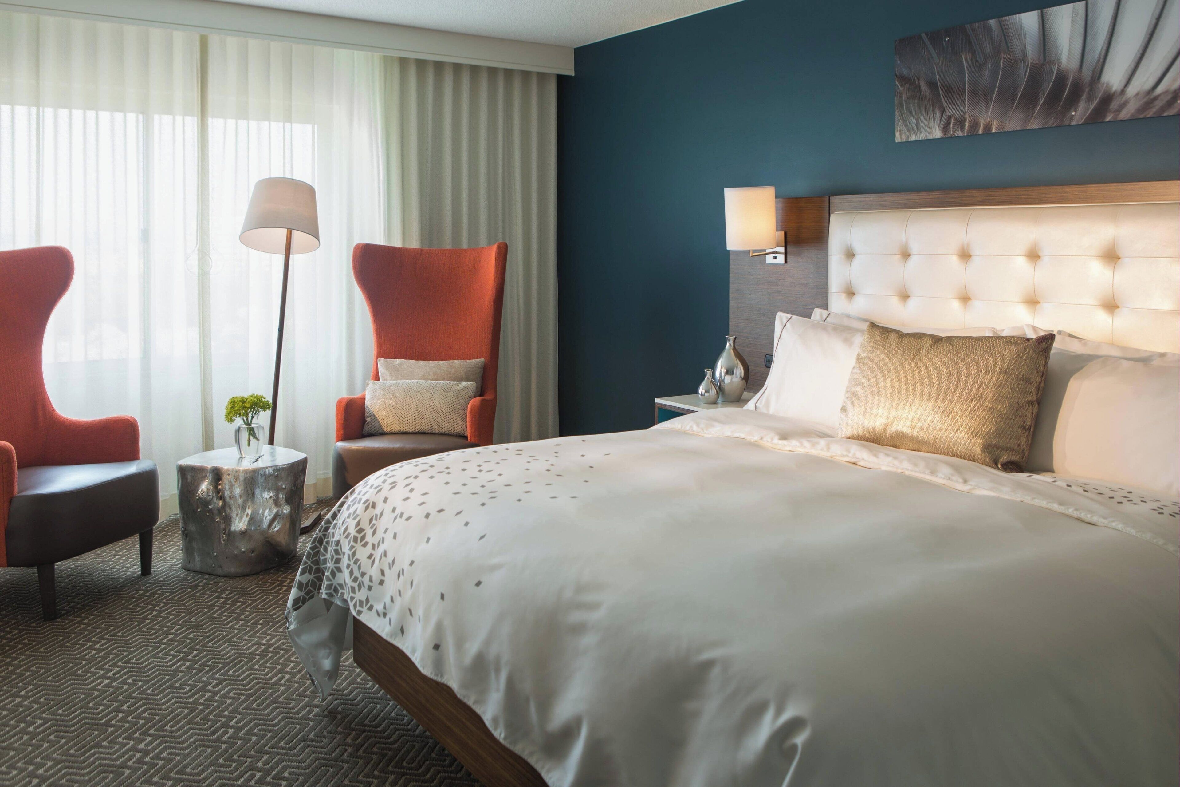 A hotel room with a large bed, two red armchairs, a side table with a vase, a floor lamp, and a wall-mounted lamp.
