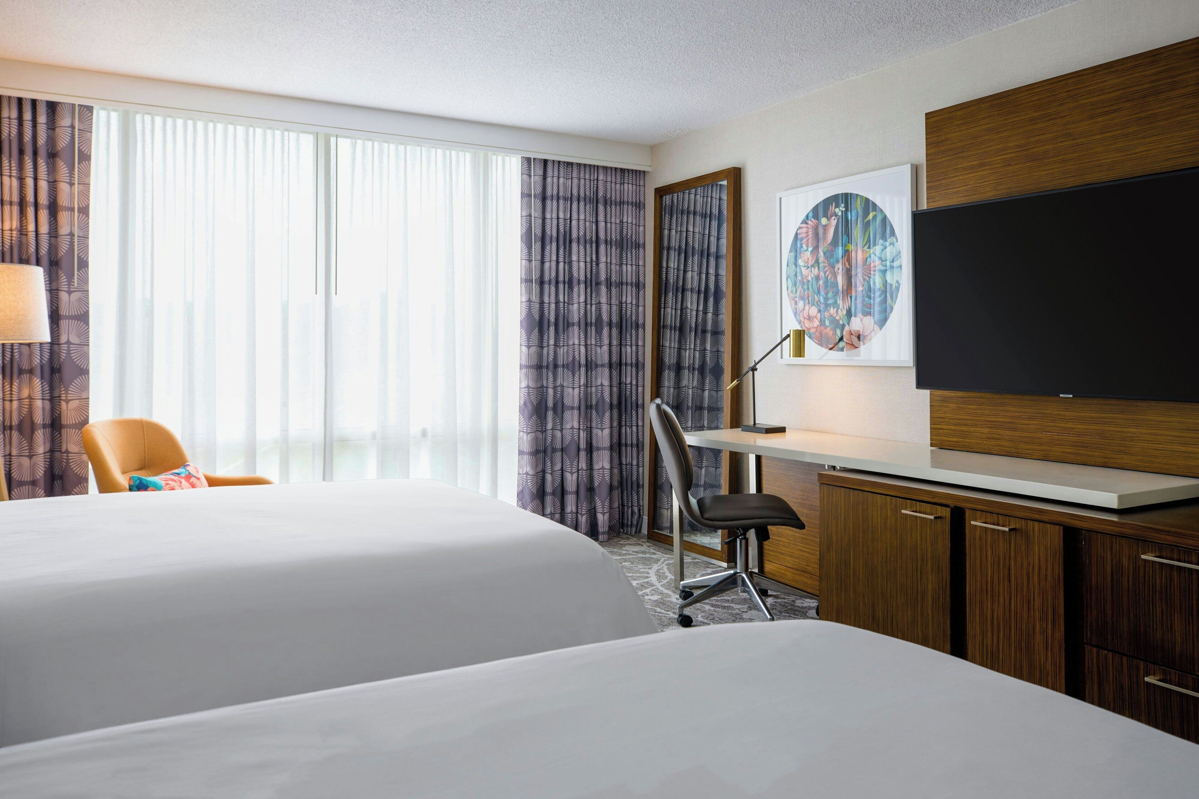 Ein Hotelzimmer mit zwei Betten, einem Schreibtisch, einem Sessel, einem Fernseher und einem großen Fenster mit Vorhängen.