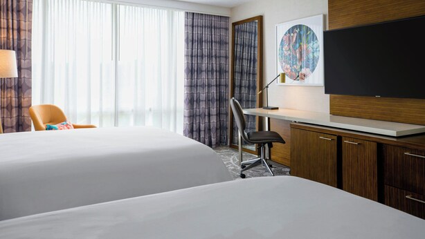 Une chambre d’hôtel avec deux lits, un bureau, une chaise, une télévision et une grande fenêtre avec des rideaux.