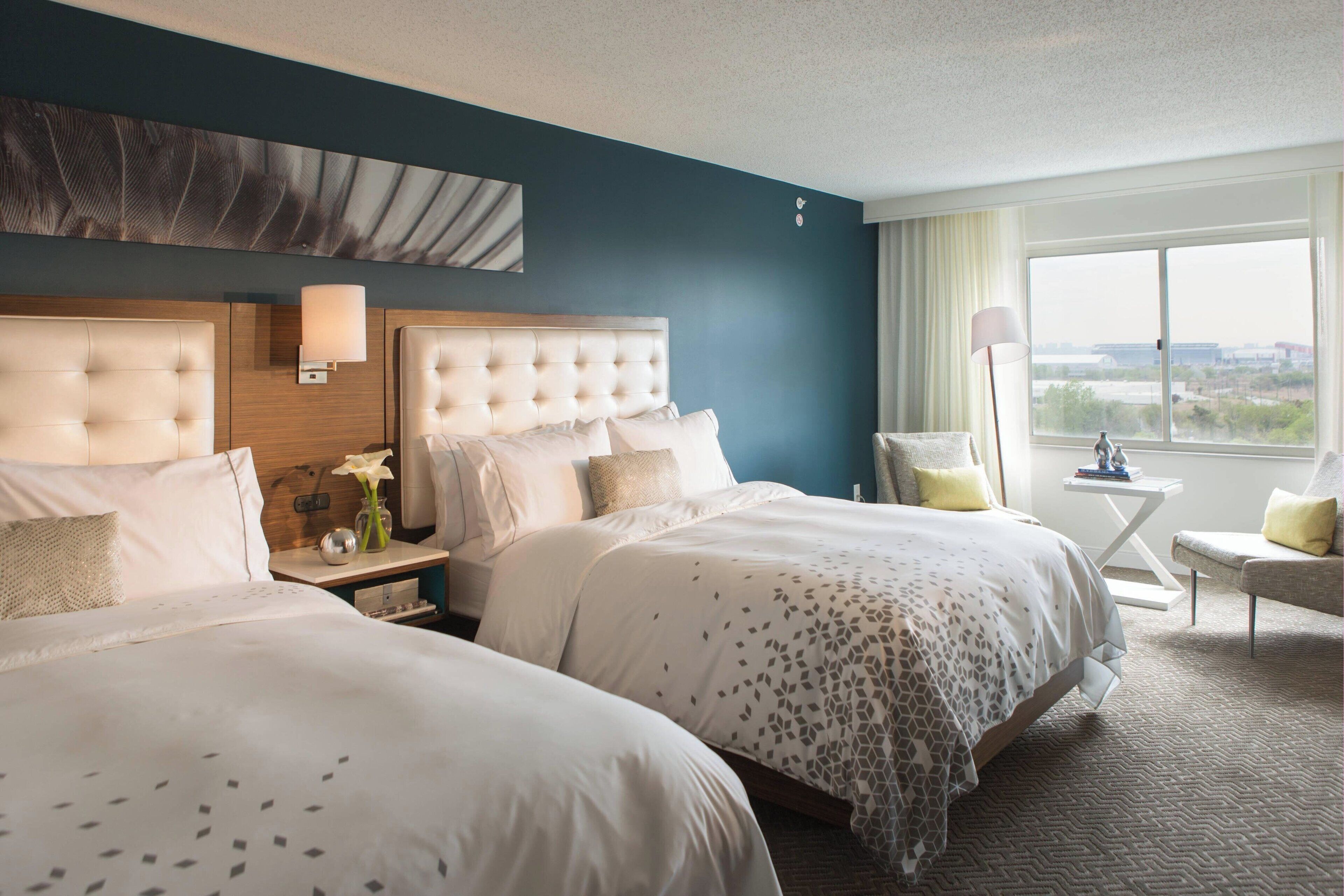 A hotel room with two beds, a nightstand, a chair, a lamp, and a view of the city.