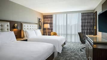 Quarto de hotel com duas camas, uma escrivaninha, uma cadeira, televisão e uma janela grande com cortinas.
