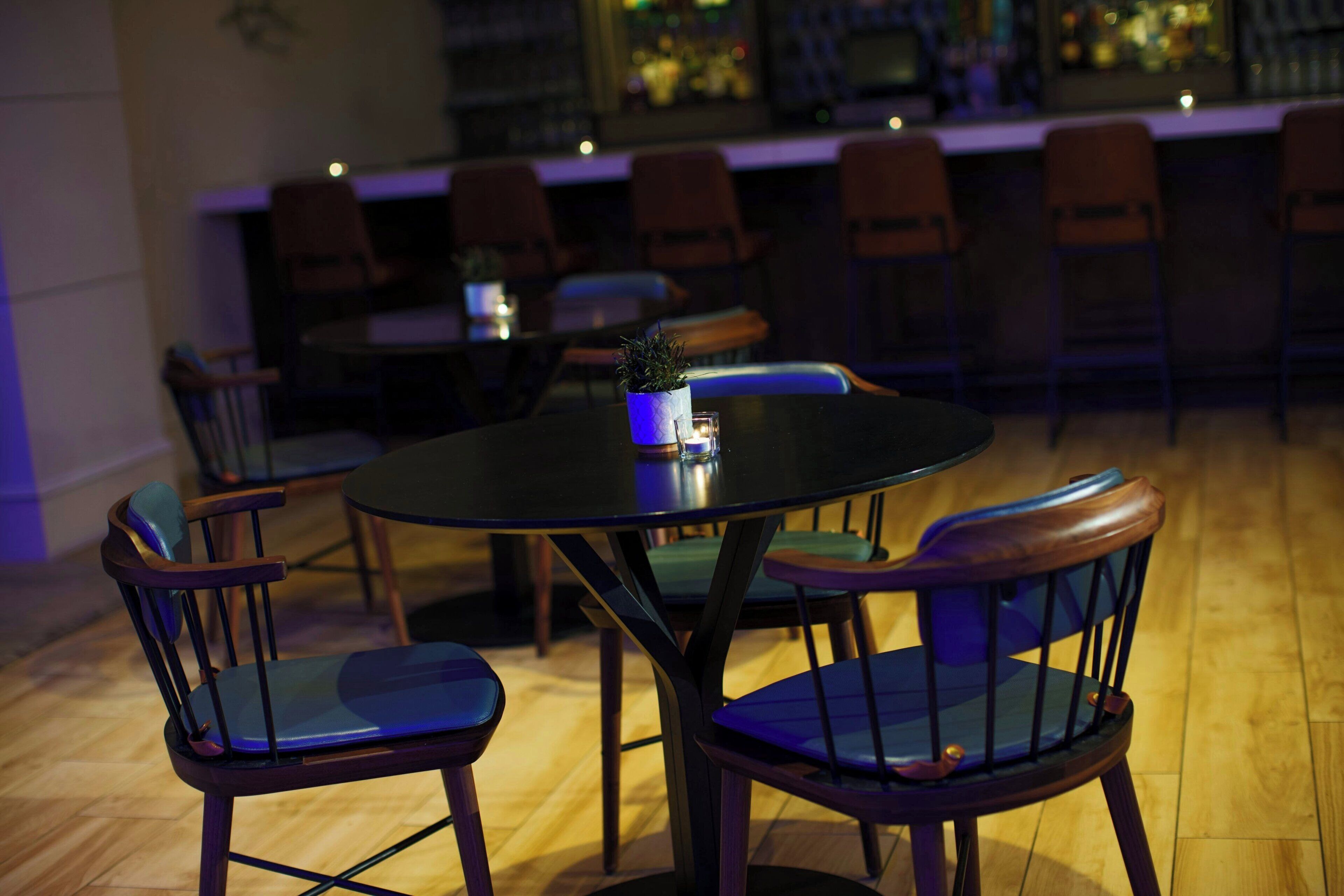 A restaurant interior with round tables and wooden chairs, a small potted plant on one table, and a bar in the background.