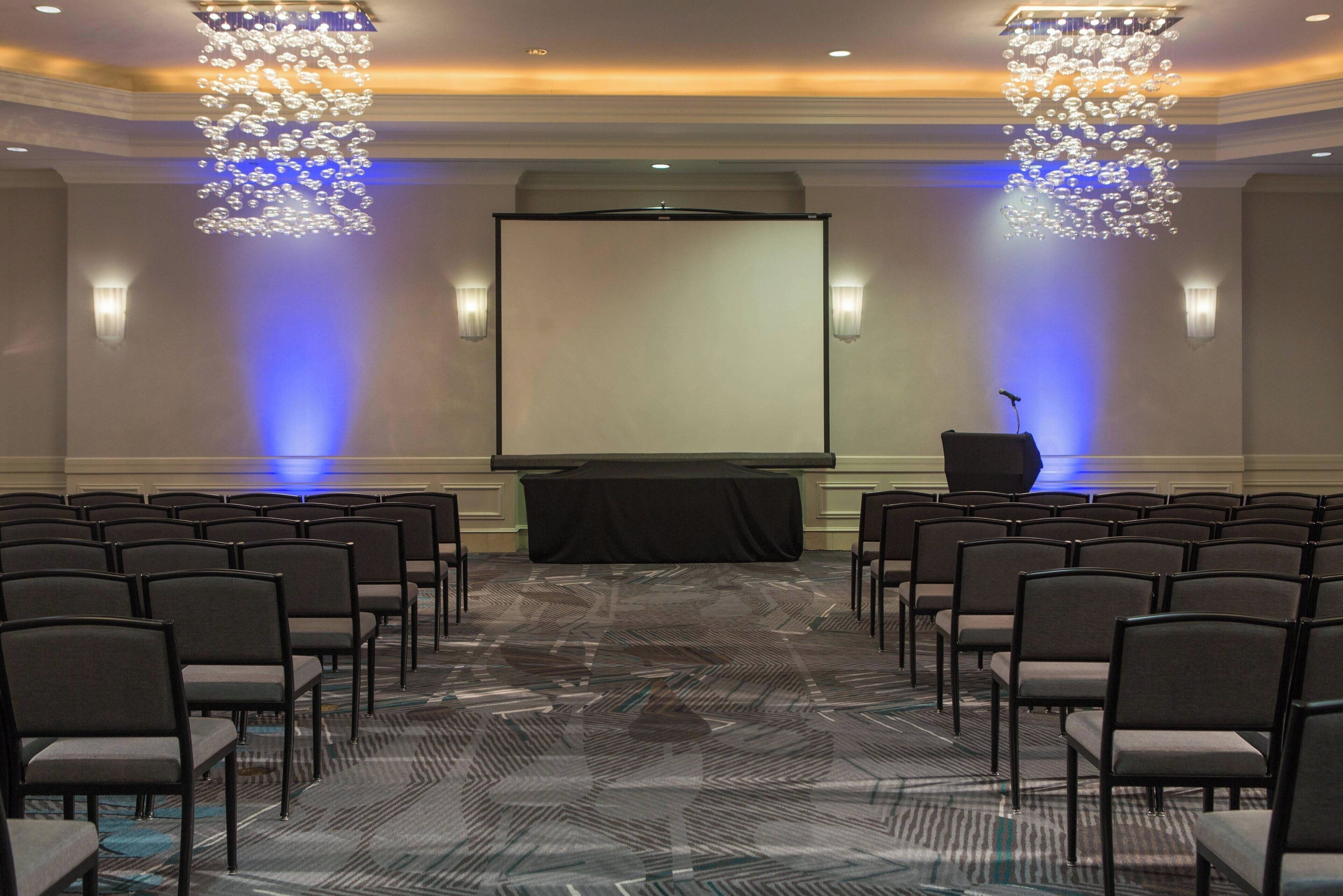 A conference room with rows of chairs, a podium, and a large screen.
