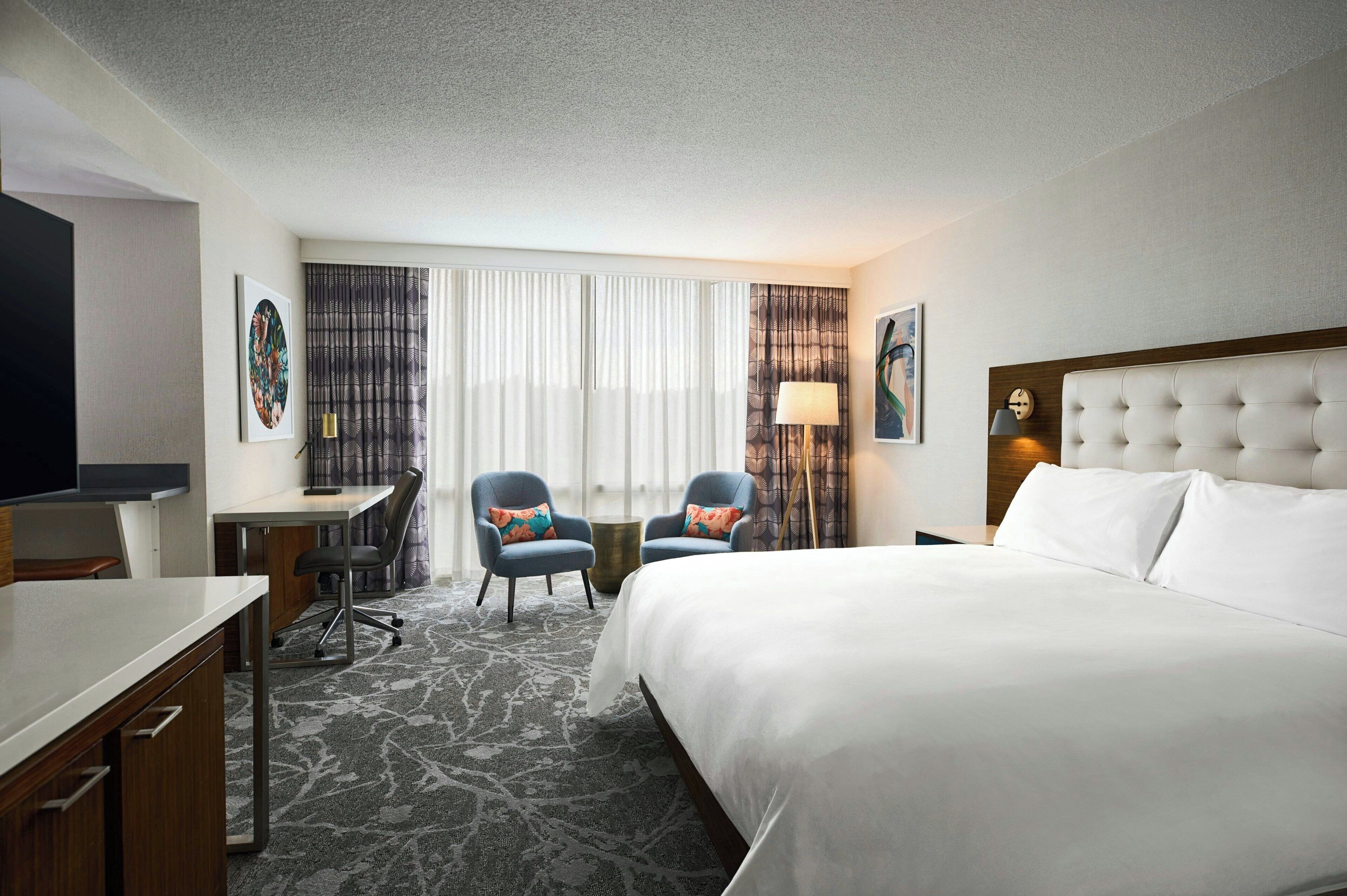 A hotel room with a large bed, a desk with a chair, two armchairs, a TV, and a window with curtains.