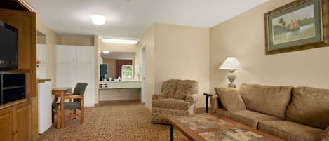 Una sala de estar con sofá, sillón y mesa de centro. Hay un televisor, una lámpara y un cuadro en la pared.