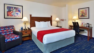 A hotel room with a large bed, two bedside tables with lamps, a desk with a chair, a patterned armchair, and two framed artworks on the walls.