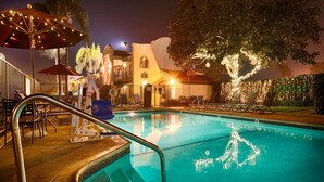 Une piscine entourée de chaises longues et de parasols, bordée d’un bâtiment à la façade blanche et d’un arbre décoré de guirlandes lumineuses.