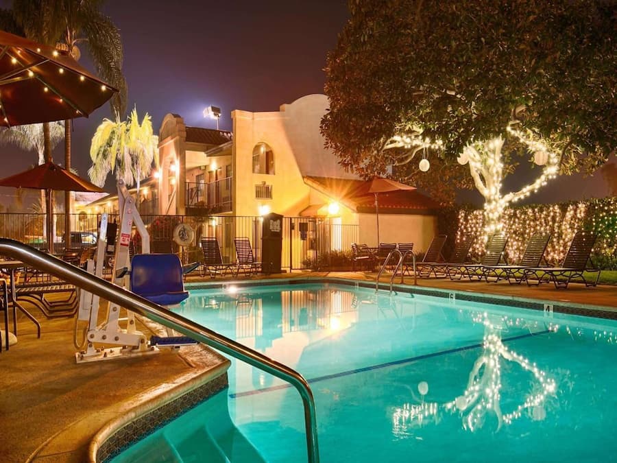 Uma piscina com espreguiçadeiras e guarda-sóis, rodeada por um edifício com fachada branca e uma árvore com luzes.