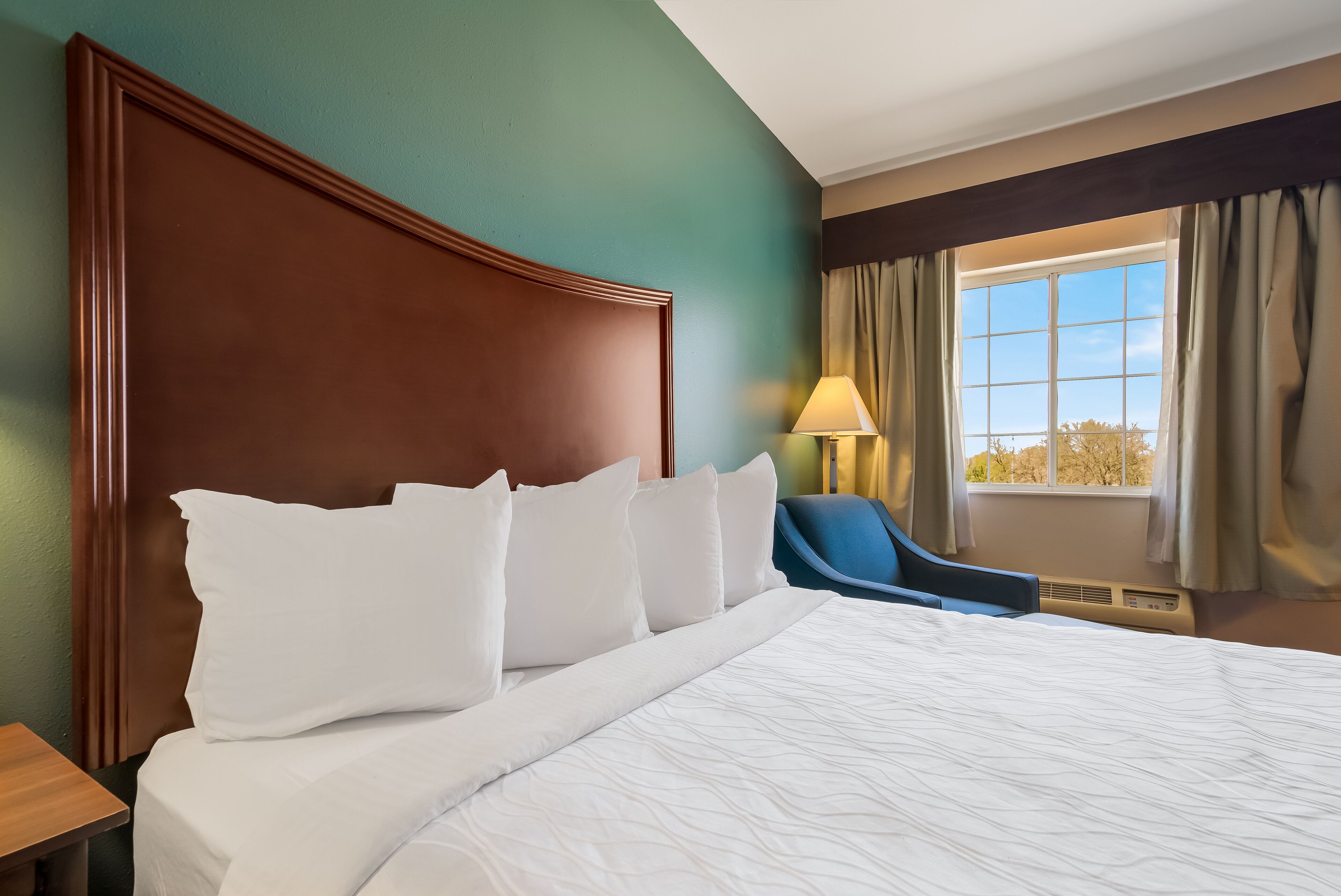 A hotel room with a large bed, a headboard, a desk, a chair, a window with curtains, and a lamp.