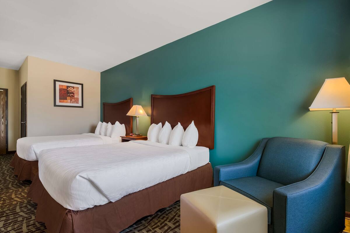 A hotel room with two beds, a blue armchair, a small table, and a lamp.