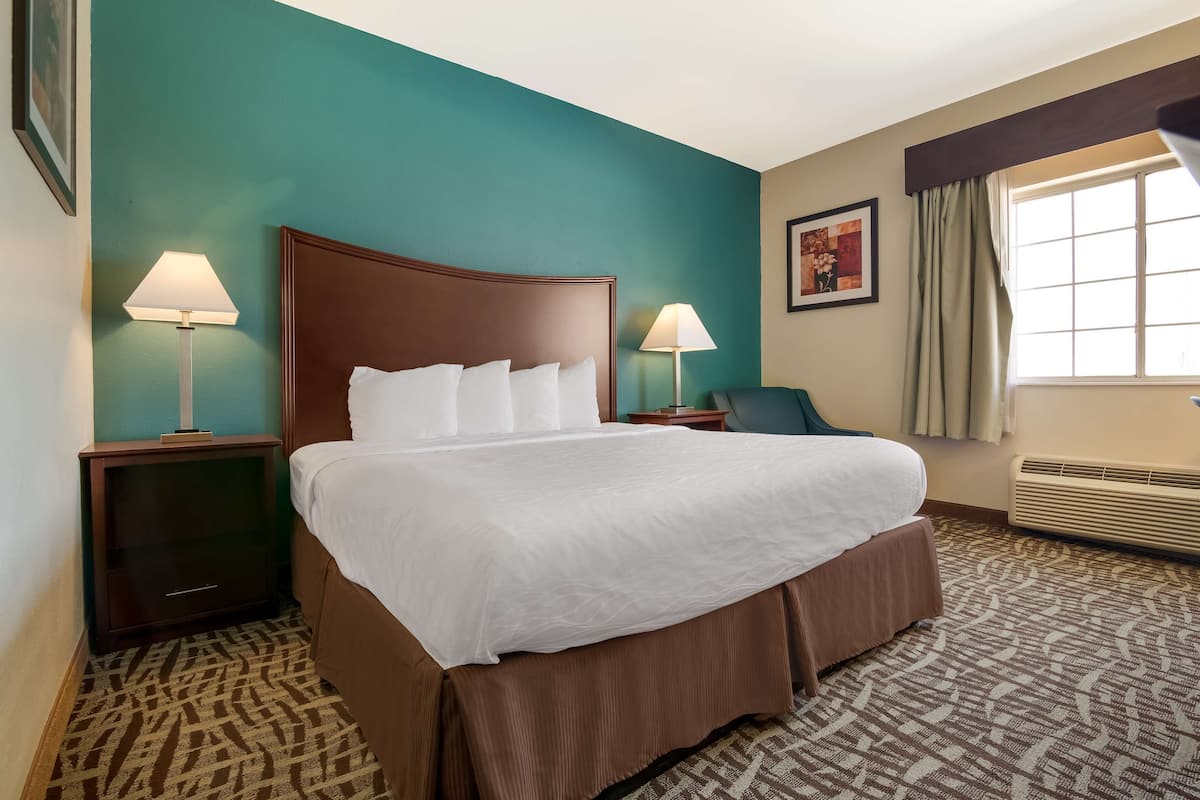 A hotel room with a large bed, two bedside tables with lamps, a chair, a window with curtains, and a wall-mounted air conditioner.