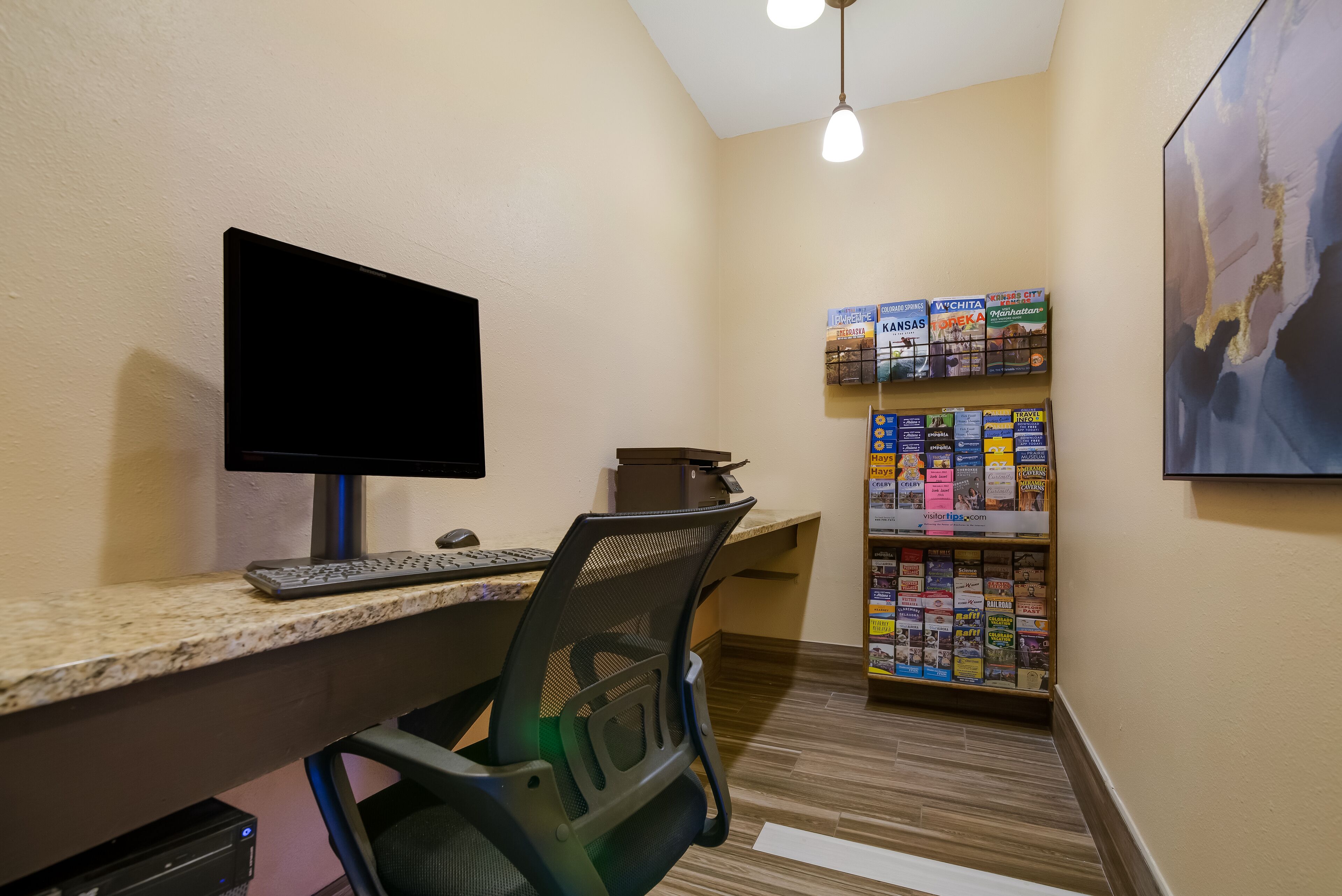 Quarto de hotel com secretária, computador, cadeira e um painel com folhetos de viagem.