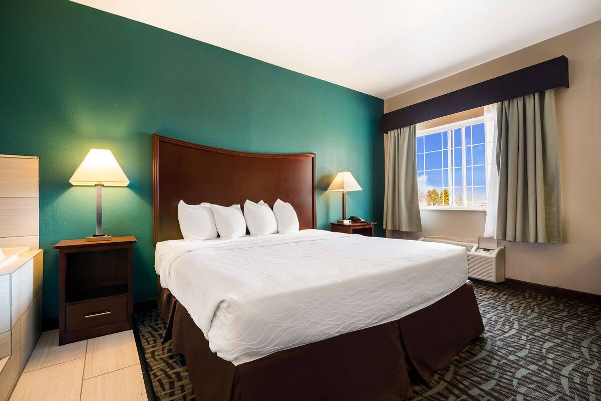 A hotel room with a large bed, two bedside tables with lamps, a window with curtains, and a teal-colored wall.