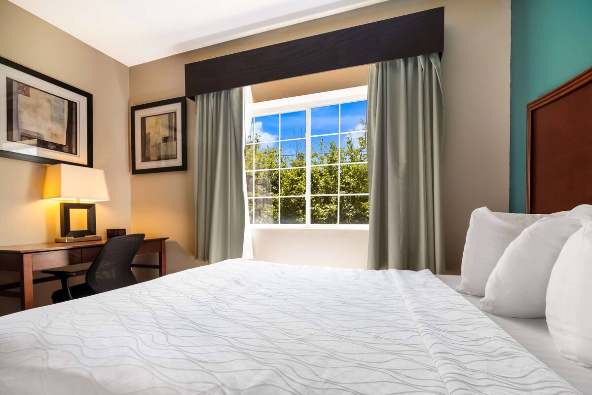 A hotel room with a large bed, a desk with a chair, a window with curtains, and framed artwork on the wall.