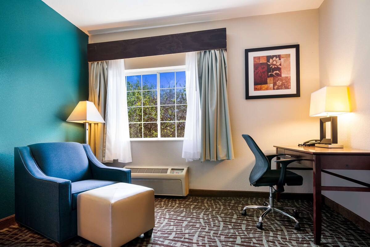A hotel room with a blue armchair, a white ottoman, a desk with a lamp, and a window with curtains.