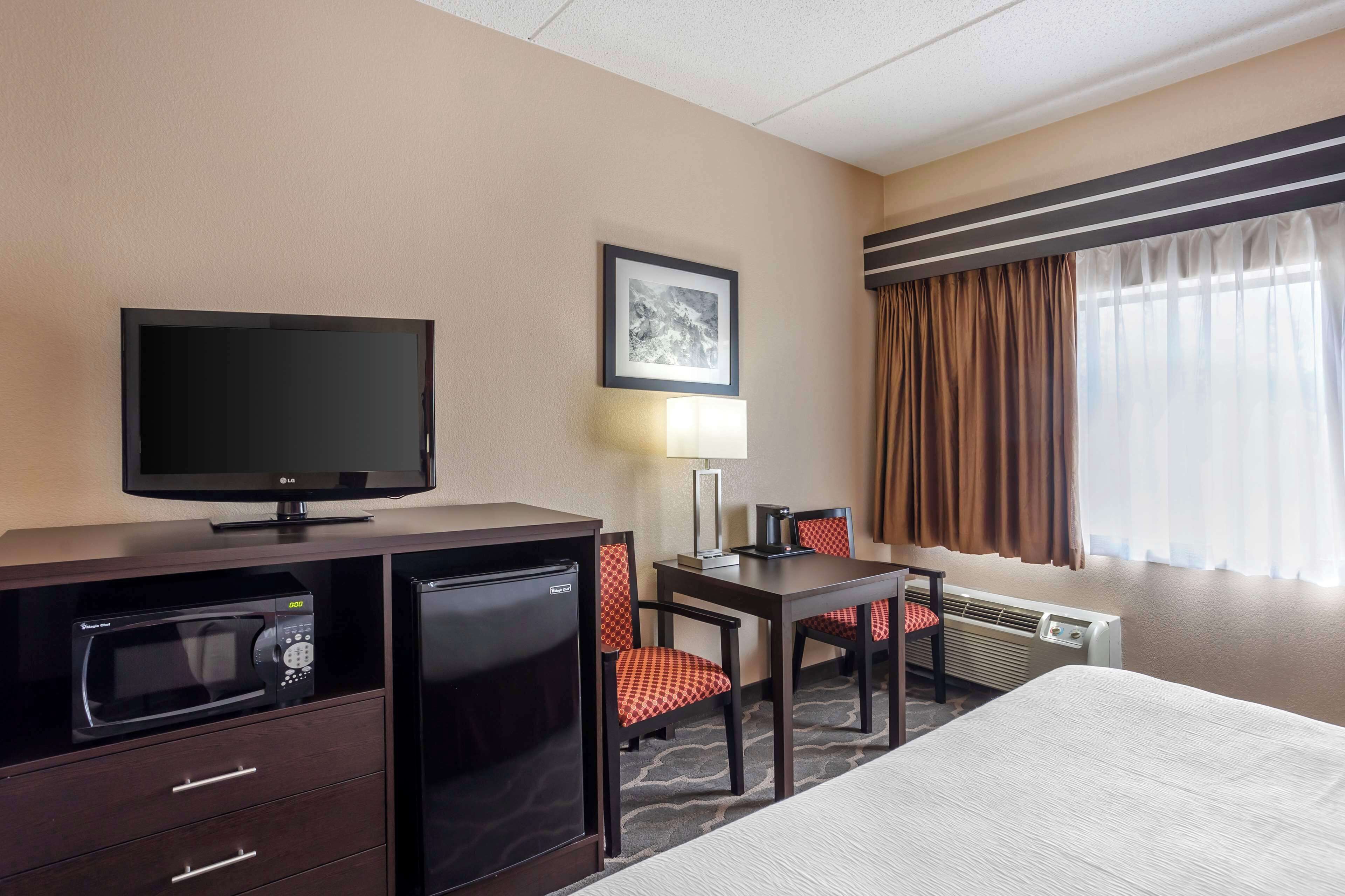 Quarto de hotel com cama, TV, micro-ondas, frigorífico, secretária com cadeira, candeeiro e janela com cortinas.