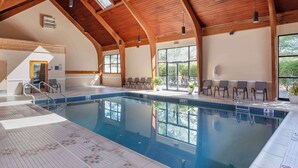 Una piscina interior con suelo de baldosas, vigas de madera en el techo y grandes ventanales.