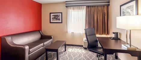 Habitación de hotel con sofá de cuero marrón, una mesita, un escritorio con una silla, una lámpara y un cuadro en la pared.