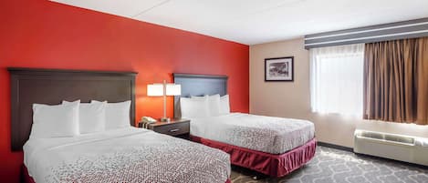 A hotel room with two beds, a red accent wall, a patterned carpet, a nightstand with a lamp, and a window with curtains.